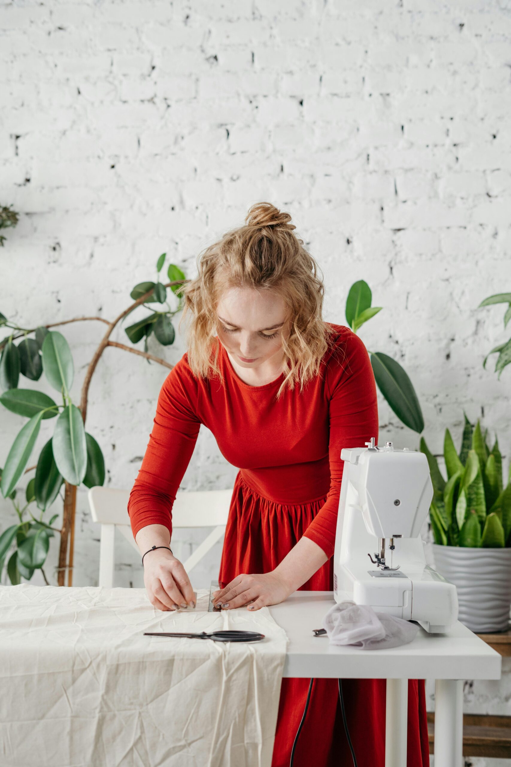 Woman in red dress sewing a garment at home with green plants around.