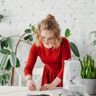 Woman in red dress sewing a garment at home with green plants around.